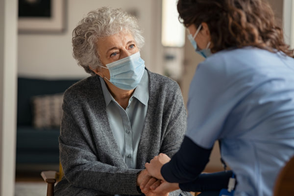 worried elderly woman talking and holding hands with female doctor while wearing face protective mask